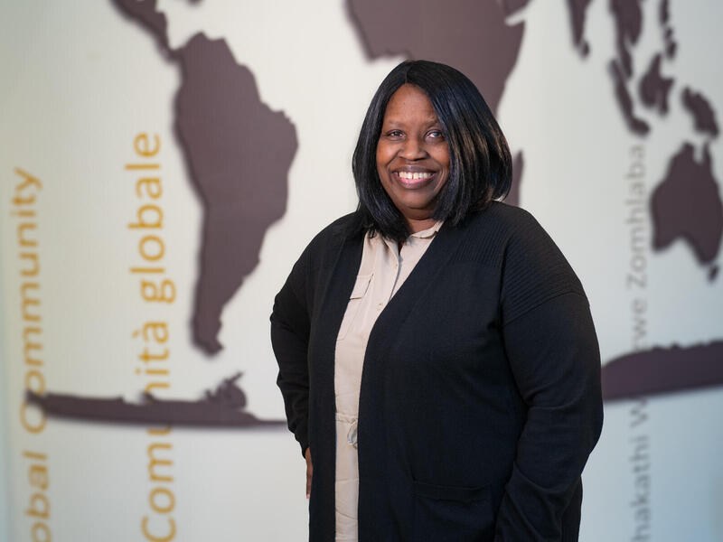 A photo of a woman standing from the waist up. Behind her is a mural of the world map. 