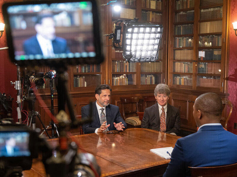 Two men sit on one side of a table in a library while a journalist sits across from them with his back to the camera. A light sits behind the men being interviewed, while a video camera in the foreground captures the image of one interviewee who is speaking.