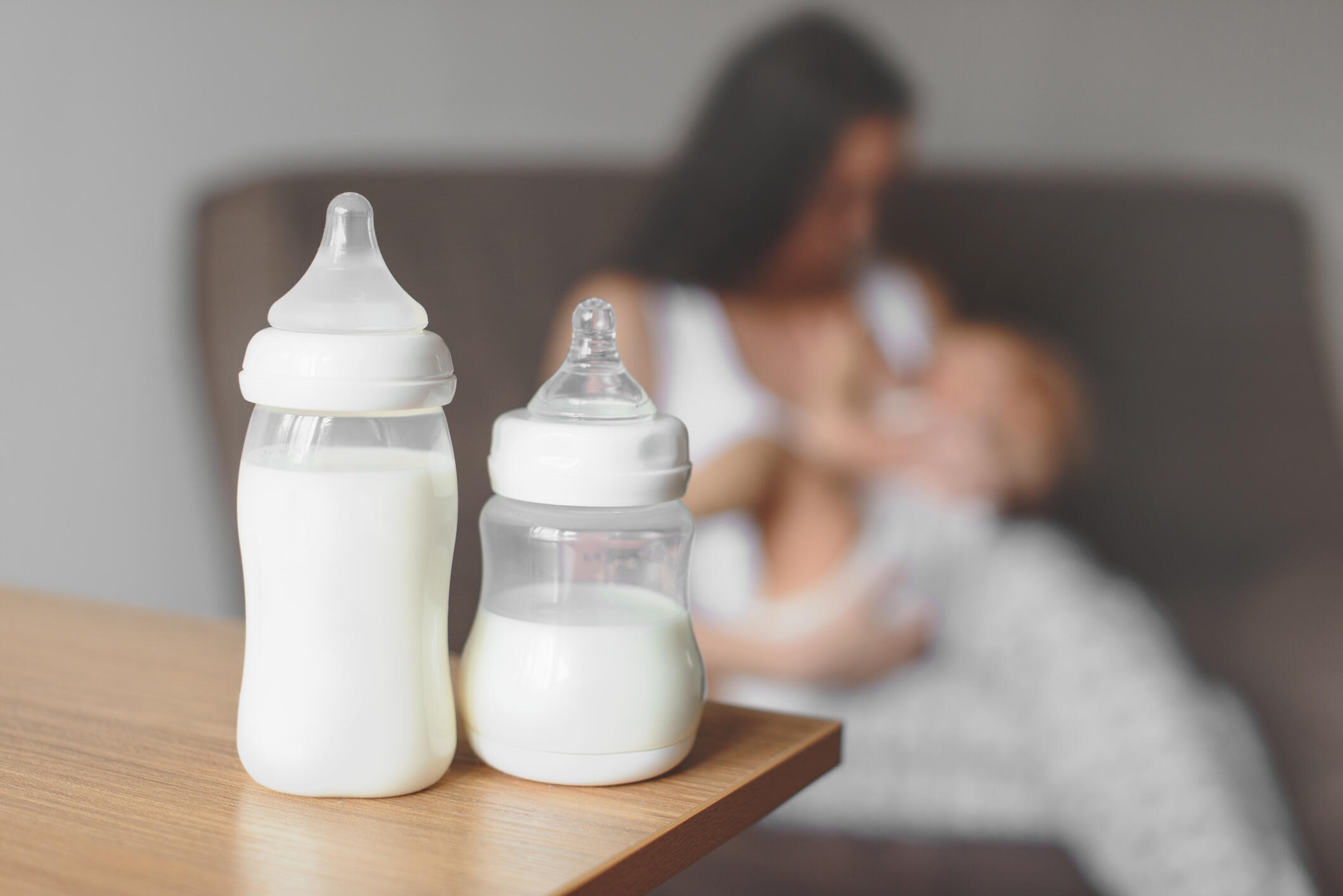 In the foreground are two bottles of formula on a table. Behind them a mother is nursing a baby in a chair. 