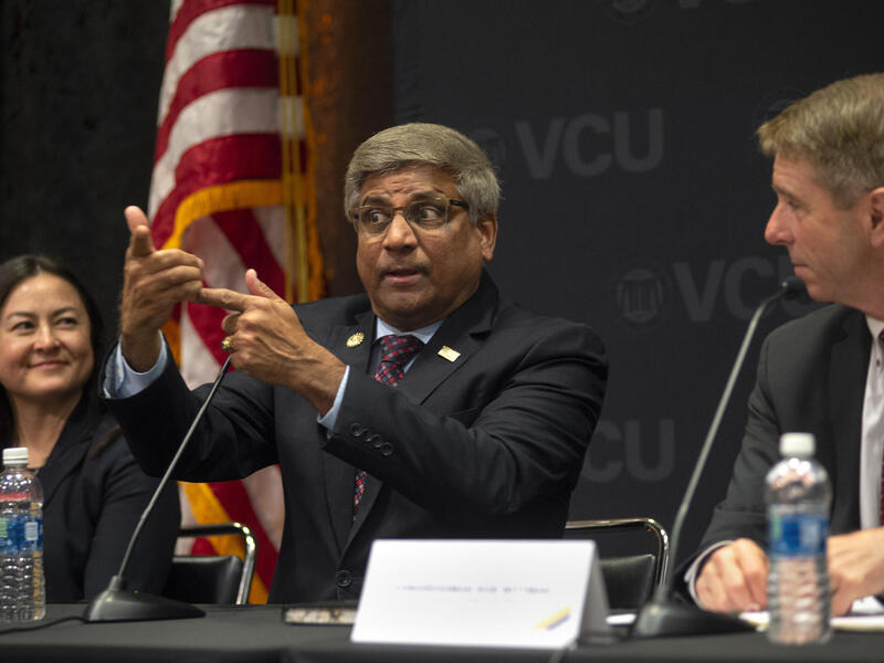 A photo of three people sitting at a table with microphones on it. The two people on the left and right are looking at a man in the middle who is talking. 