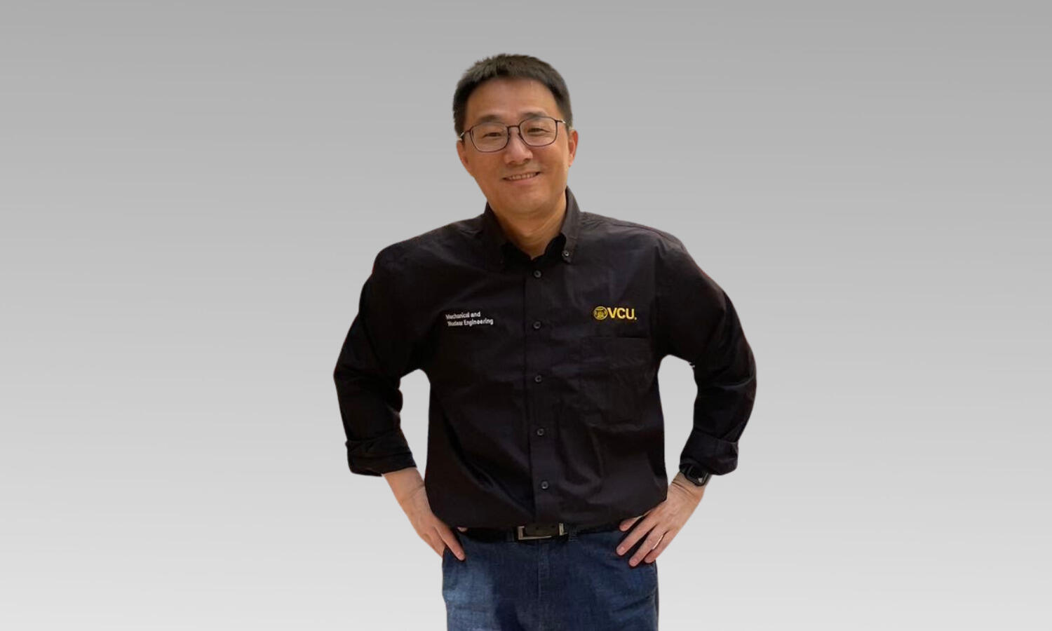 A man in a black shirt and glasses smiles with his hands on his hips.