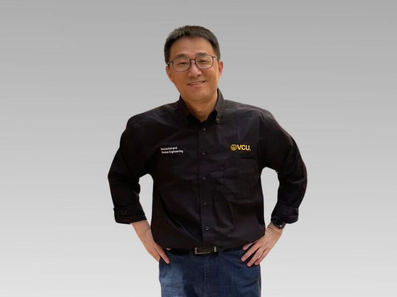 A man in a black shirt and glasses smiles with his hands on his hips.