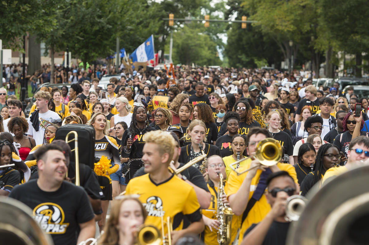 A crowd of people walking down a street. In the front of the crowd some of the people are holding instruments. 