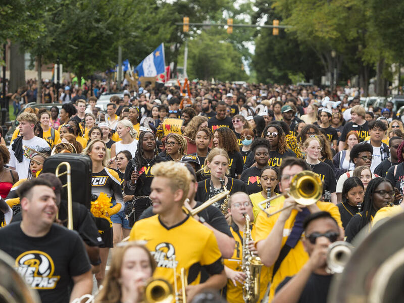 A crowd of people walking down a street. In the front of the crowd some of the people are holding instruments. 