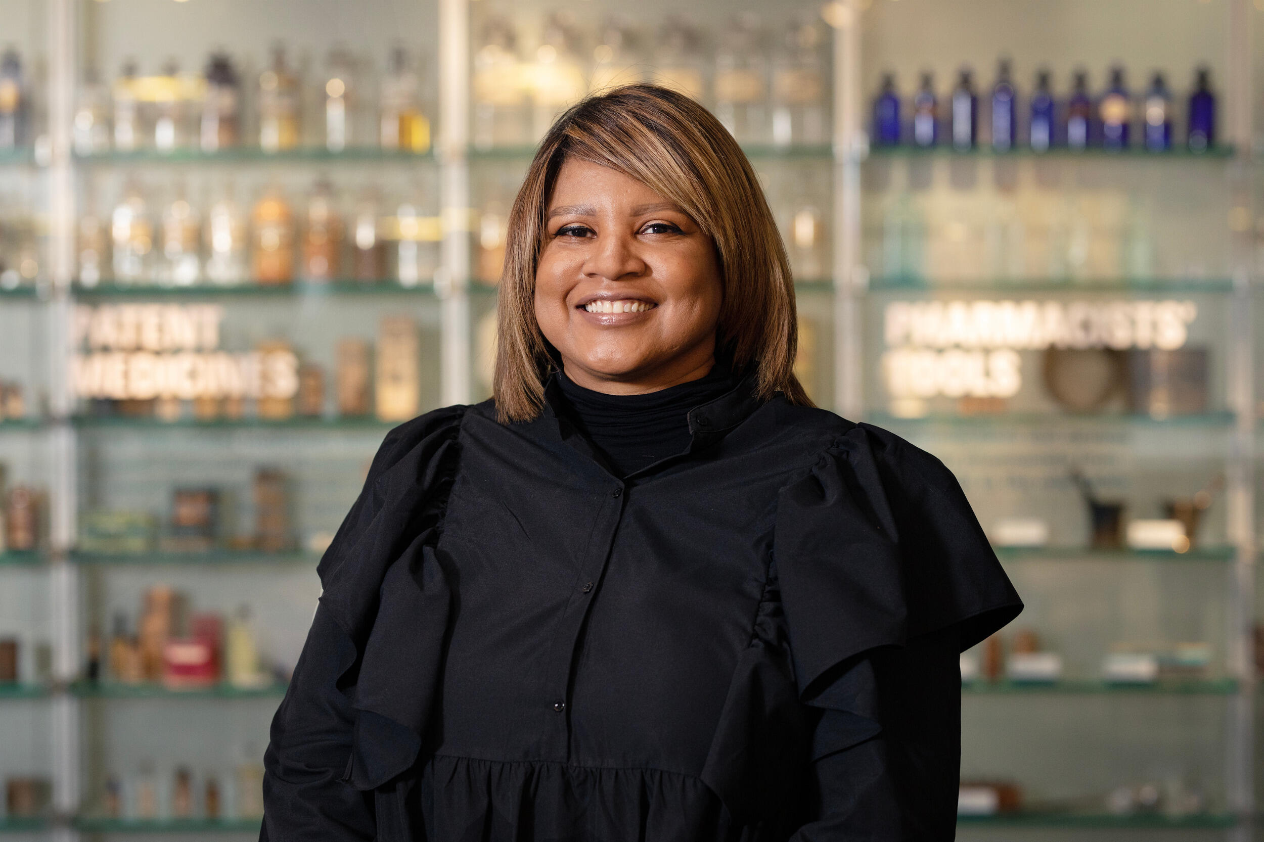 A woman wearing black smiles while standing in front of shelves holding bottles.