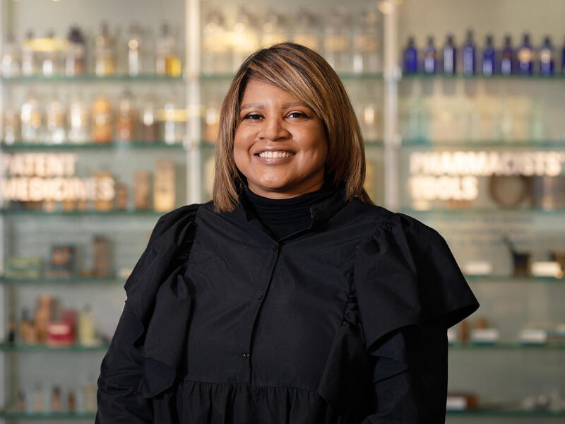 A woman wearing black smiles while standing in front of shelves holding bottles.