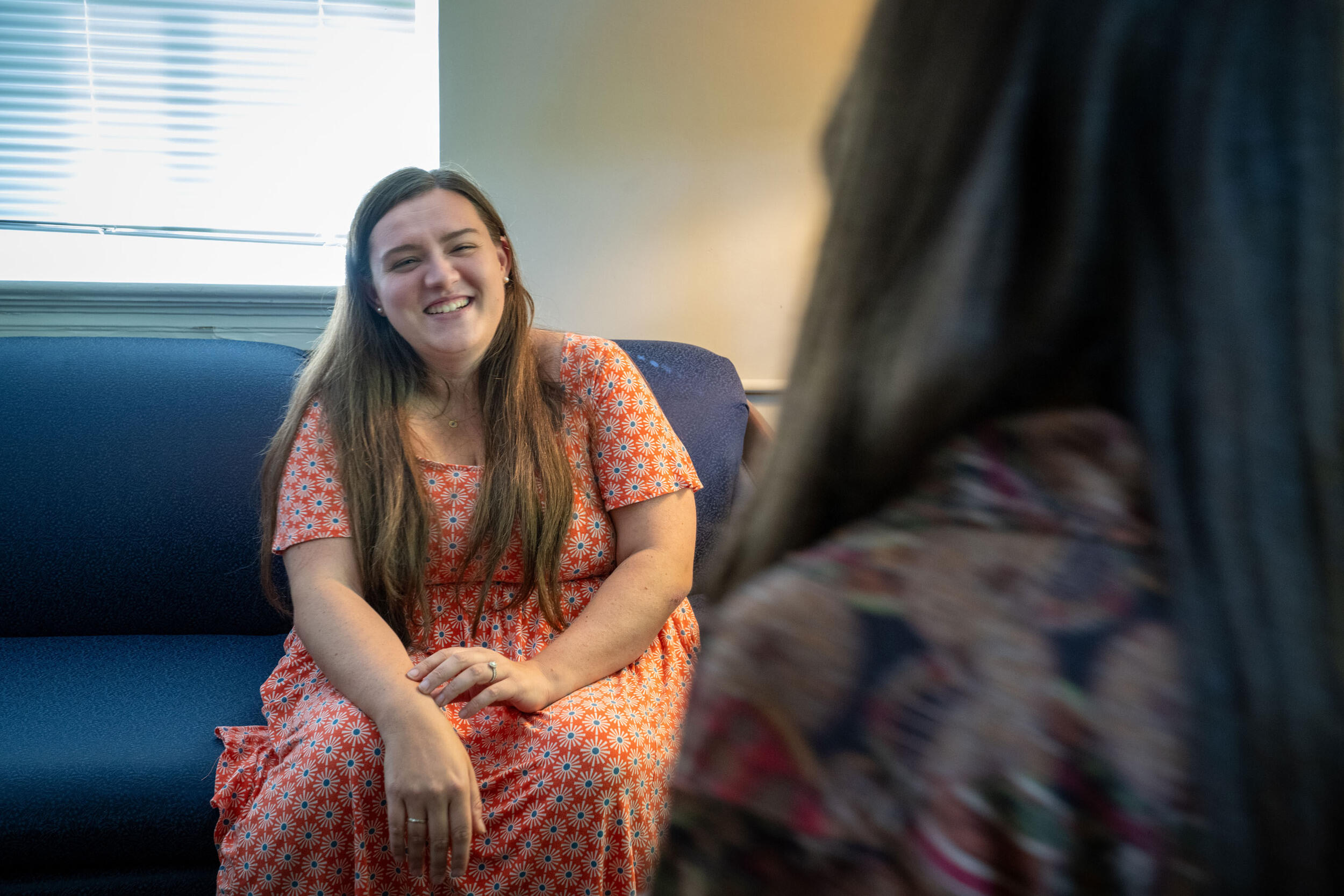 A photo of a woman sitting on a couch smiling at another person sitting across from her. 