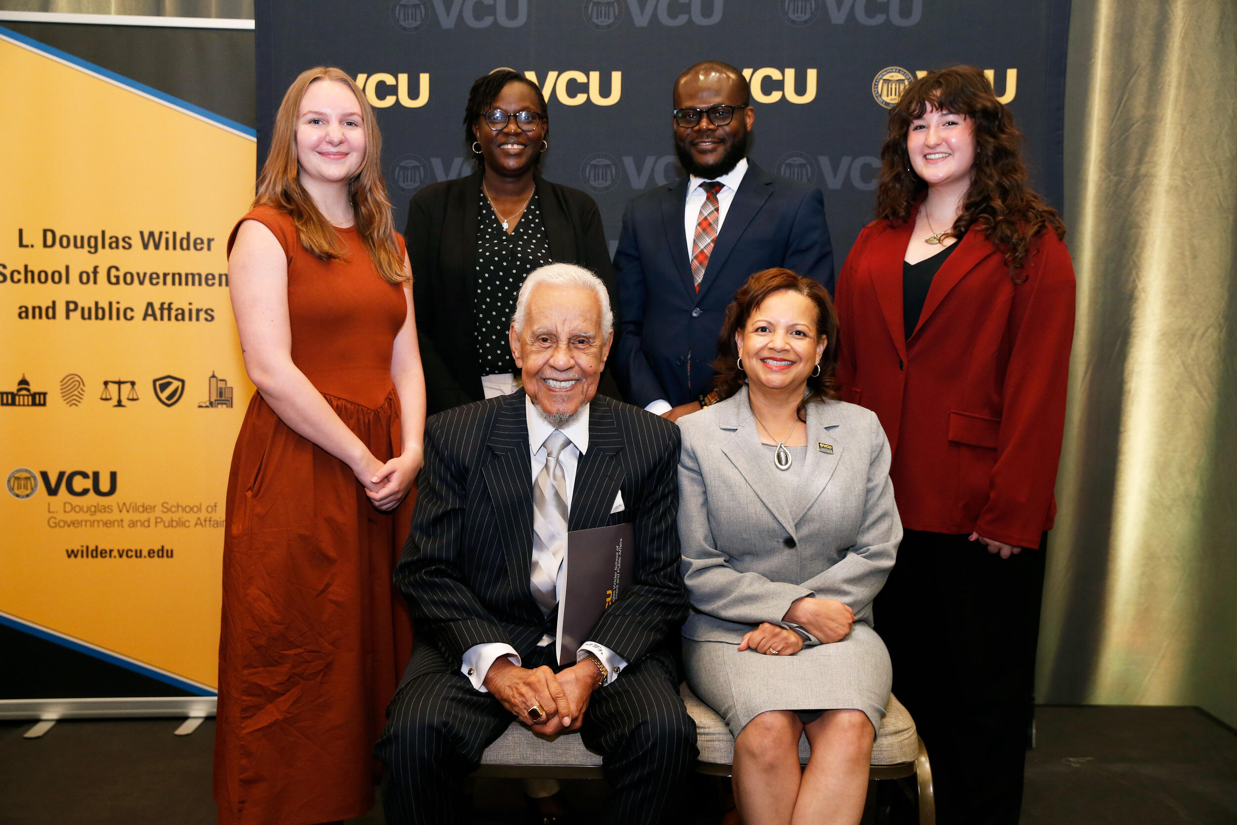 A photo of four people standing and two people sitting. 