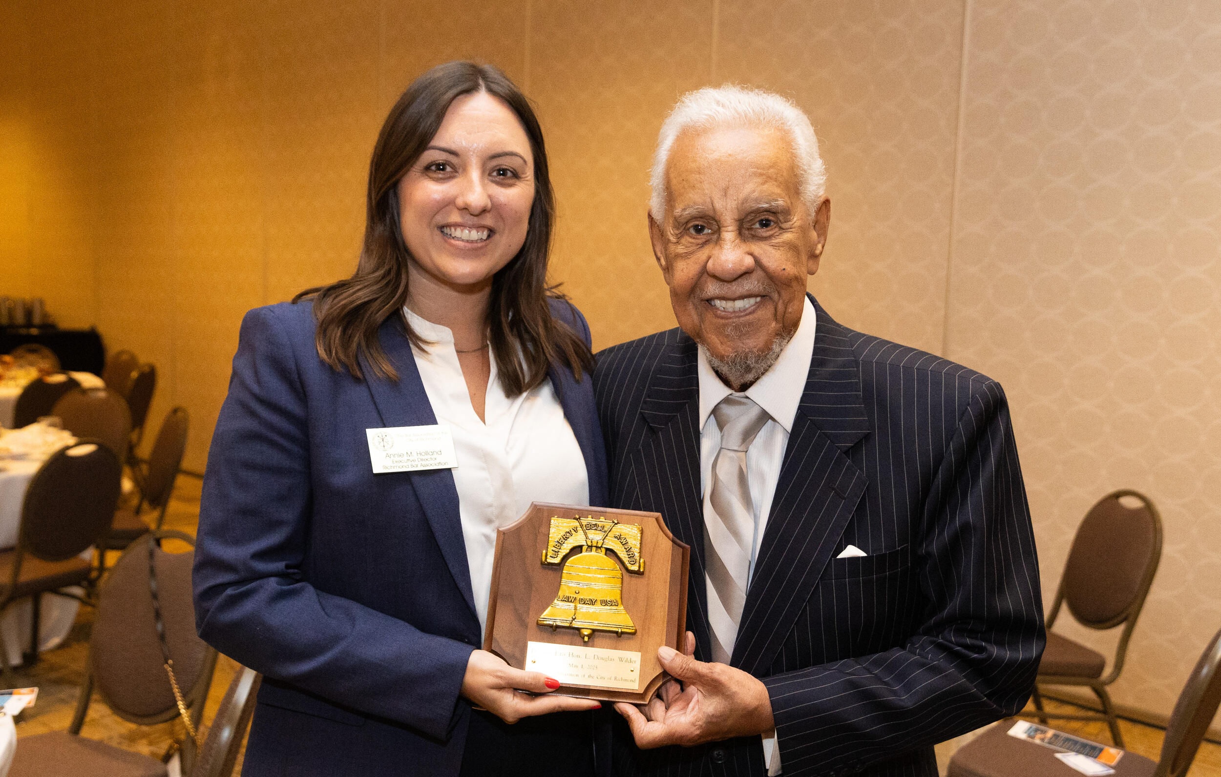 A woman and a man are standing next to each other, holding an award plaque with an illustration of a cracked bell on it. 