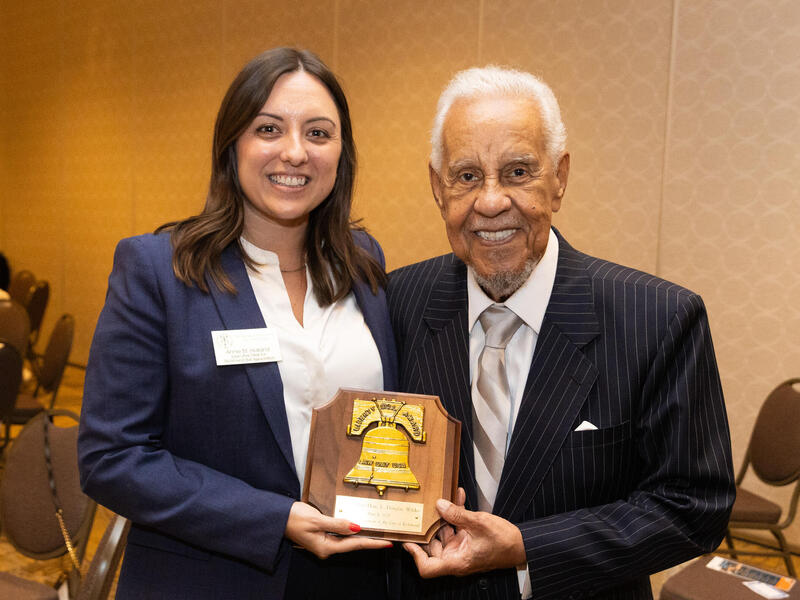 A woman and a man are standing next to each other, holding an award plaque with an illustration of a cracked bell on it. 