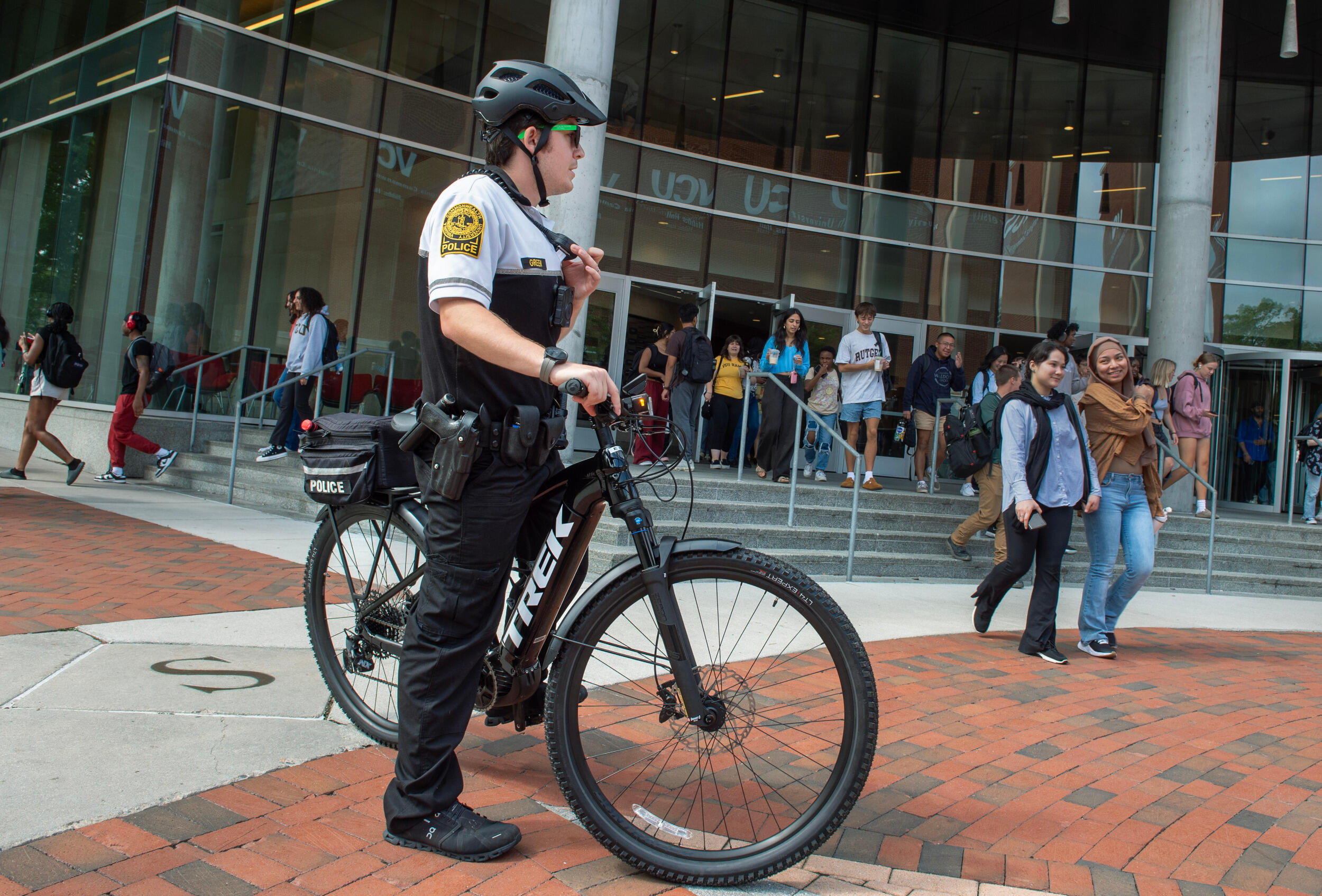 A police officer in a bike helmet stands over his bicycle on campus with students in the background.