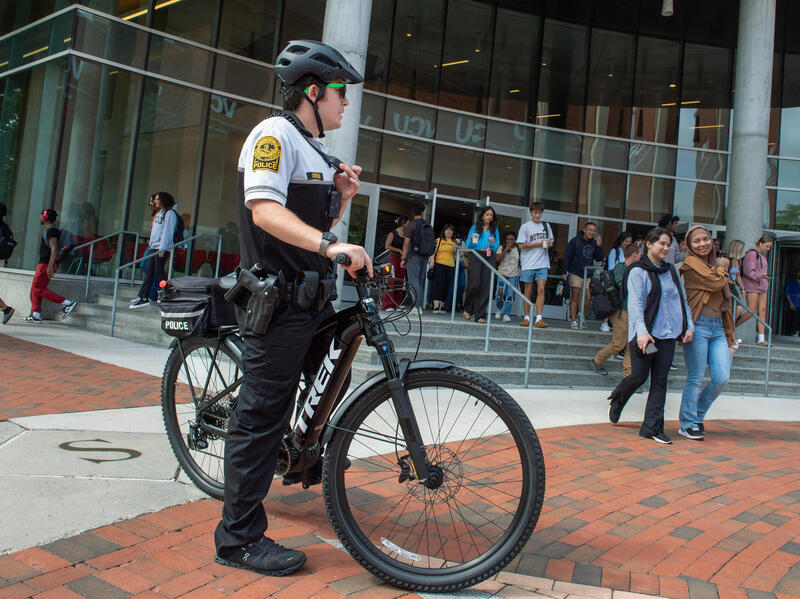 A police officer in a bike helmet stands over his bicycle on campus with students in the background.