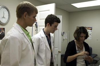 Derek Mires and William Coleman look on as Sallie Mayer demonstrates blood glucose monitoring at the CrossOver Ministry clinic on Cowardin Avenue). Image courtesy of Hal Tyler Photography.