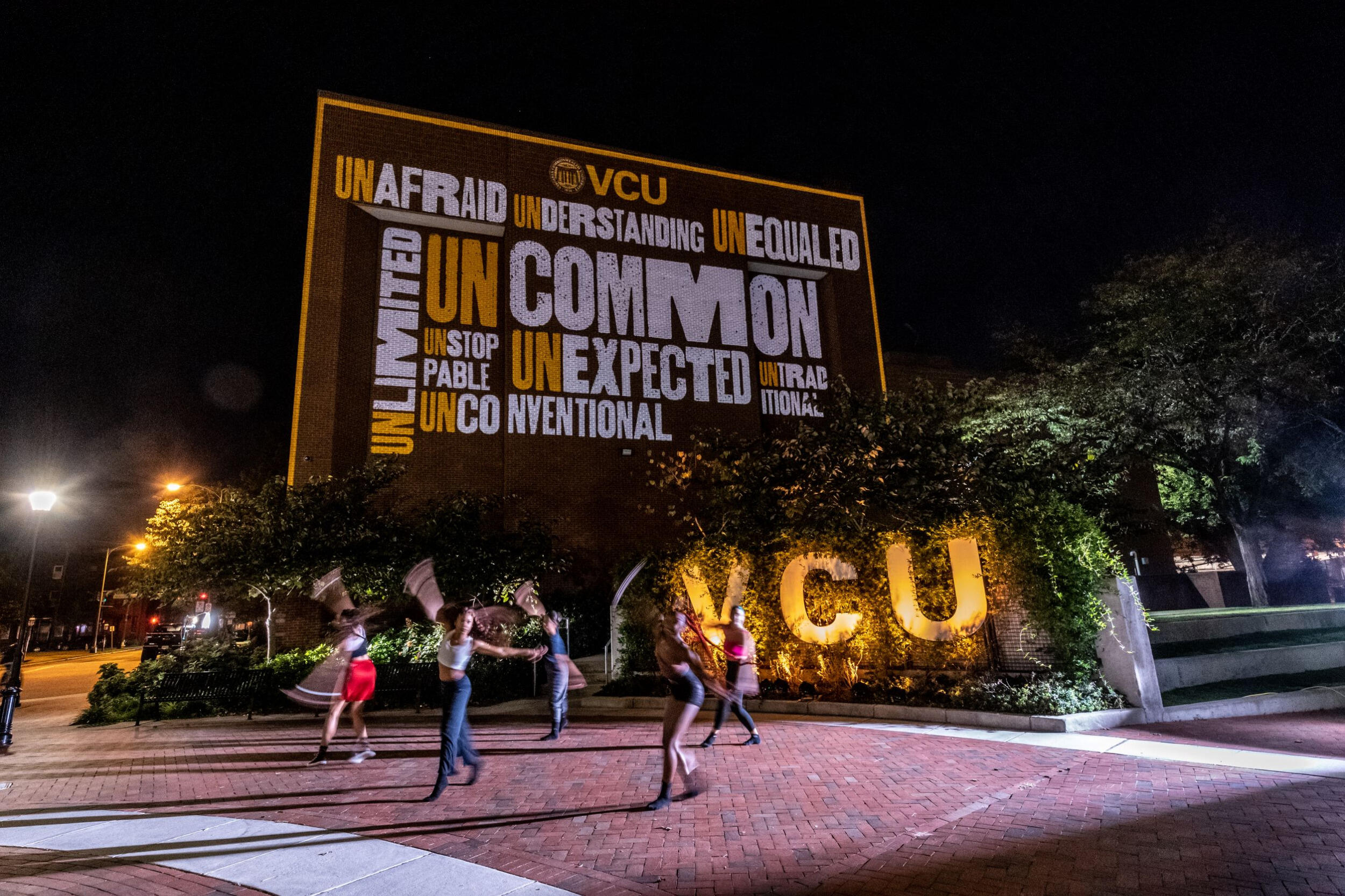 Dancers outdoors at night in front of a large sign with a variety of words that start with \"un\" and a lit VCU sign.