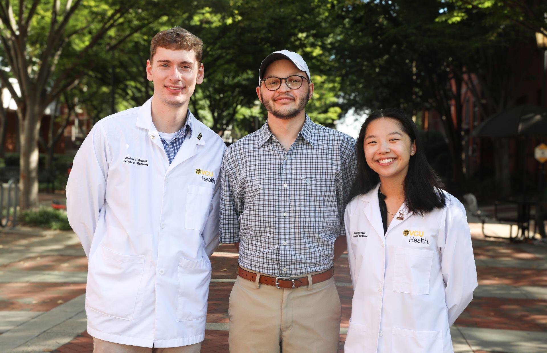 A photo of three people, two men on the left and a woman on the right. The man on the far left and the woman on the right are wearng medical coats that say \"VCU Health.\"