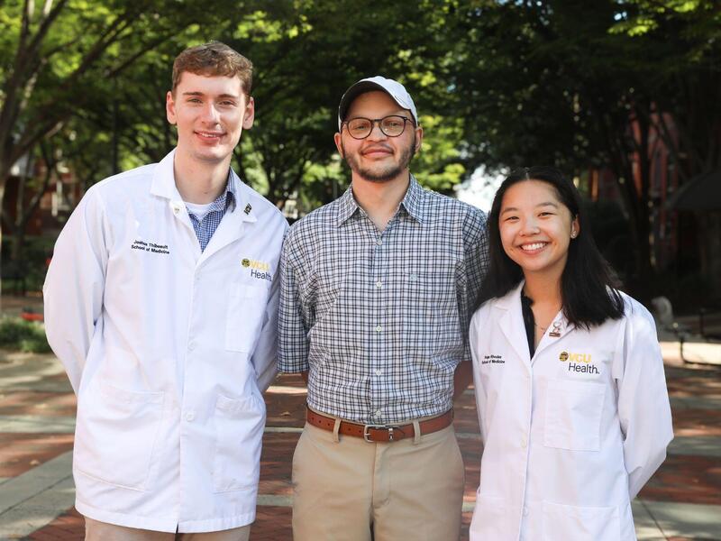 A photo of three people, two men on the left and a woman on the right. The man on the far left and the woman on the right are wearng medical coats that say \"VCU Health.\"