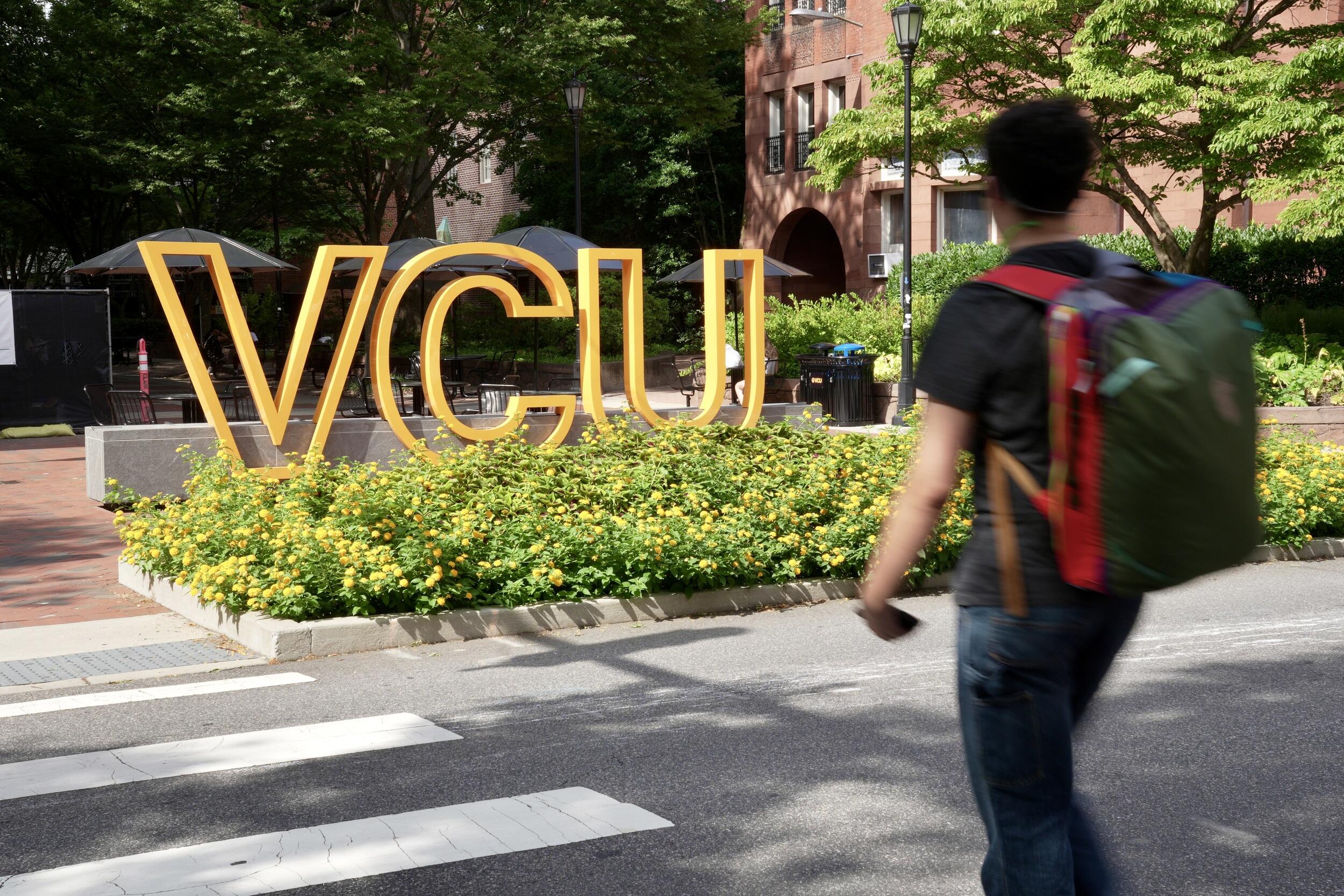 A photo of a man wearing a backpack crossing a street at a crosswalk. On the other side of the street is a sign that says \"VCU\" in large yellow letters. 