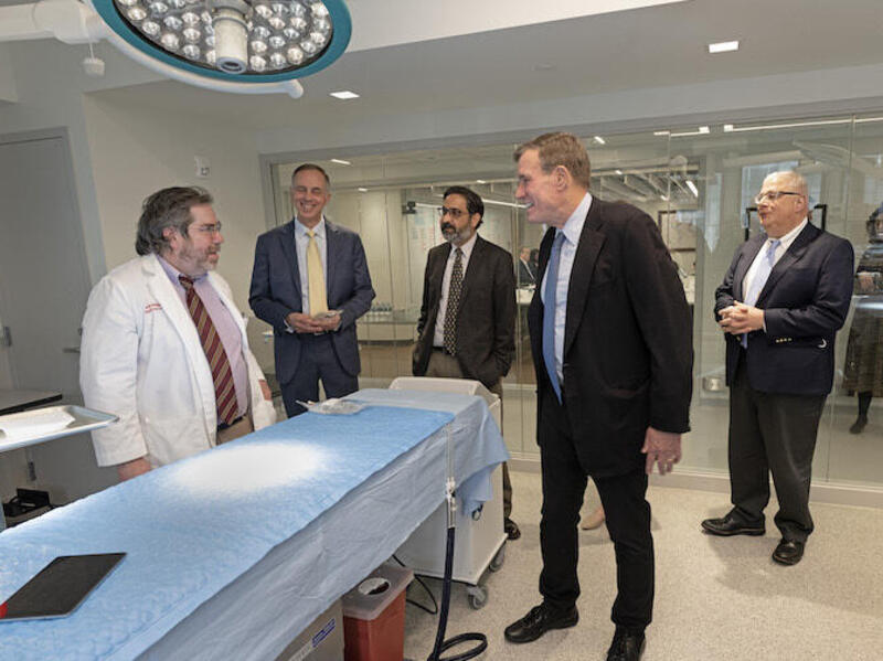 A photo of five men standing in a medical proceedure room, in a semi-circle around a medical bed. The man on the far left is wearing a white doctor's coat. The other four men are wearing suits and ties. 