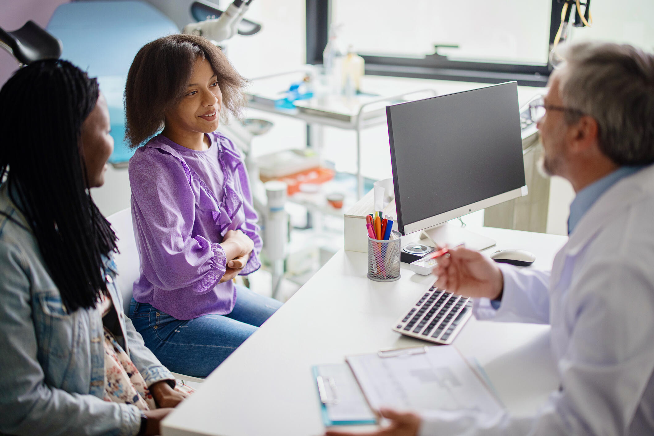 A woman and young girl sitting at a desk talking to a doctor holding a clip board and pencil. 