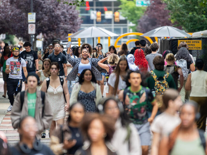 A photo of a dense crowd of people walking down a brick sidewalk. In the middle of the crowd is a woman raising up her arms and cheering as she holds her smart phone in her left hand.