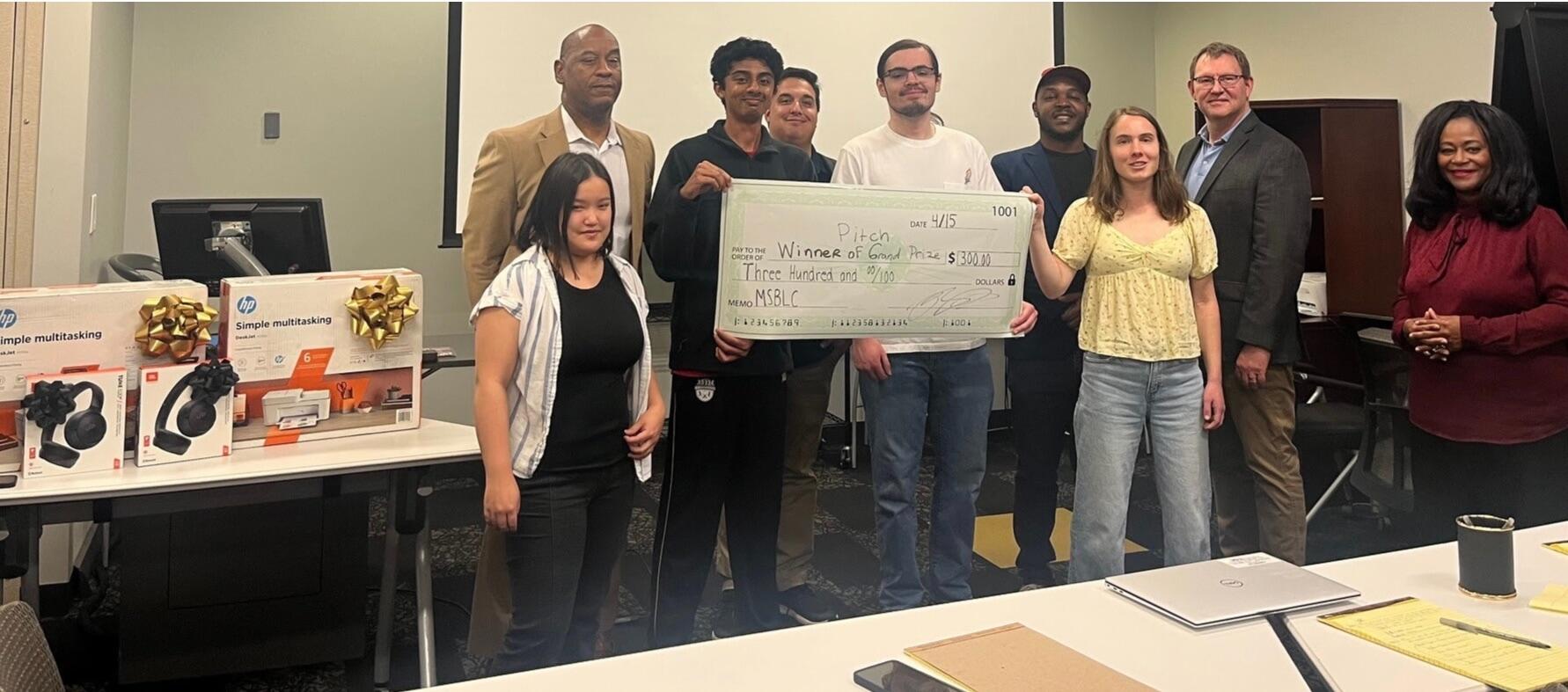 A group photo of nune people. Two men and one woman in the middle of the group are holding a giant novelty check for $300.