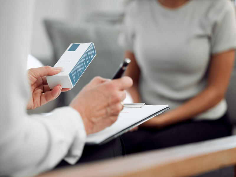A photo of a person looking at a box and writing on a clip board. Another woman is sitting across from them on a coach. 
