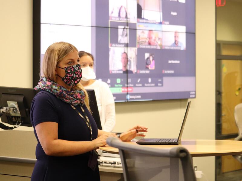 W woman wearing a face mask standing in front of a laptop. Behind her is another woman wearing a face mask. 