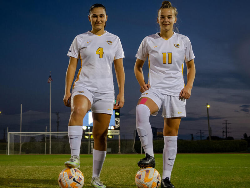 Two women wearing soccer uniforms standing on a soccer field. Each of them have one foot on top of a soccer ball. 