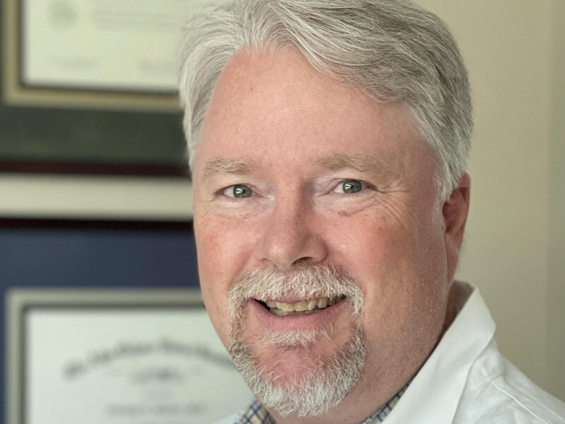 A photo of a man wearing a white medical coat. On the wall behind him are two framed degrees. 