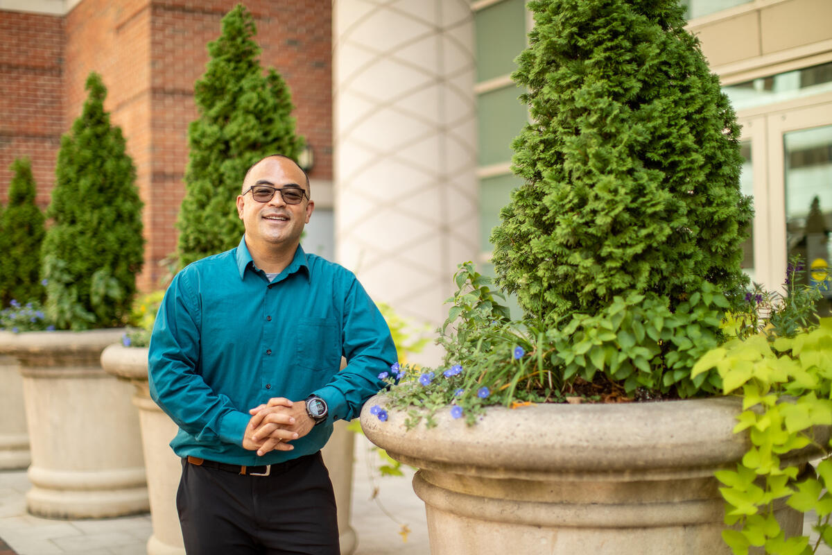 A man in a button down shirt leaning against a potted tree 