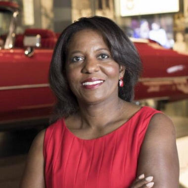 A woman wearing a red shirt standing in front of a red convertible car. 