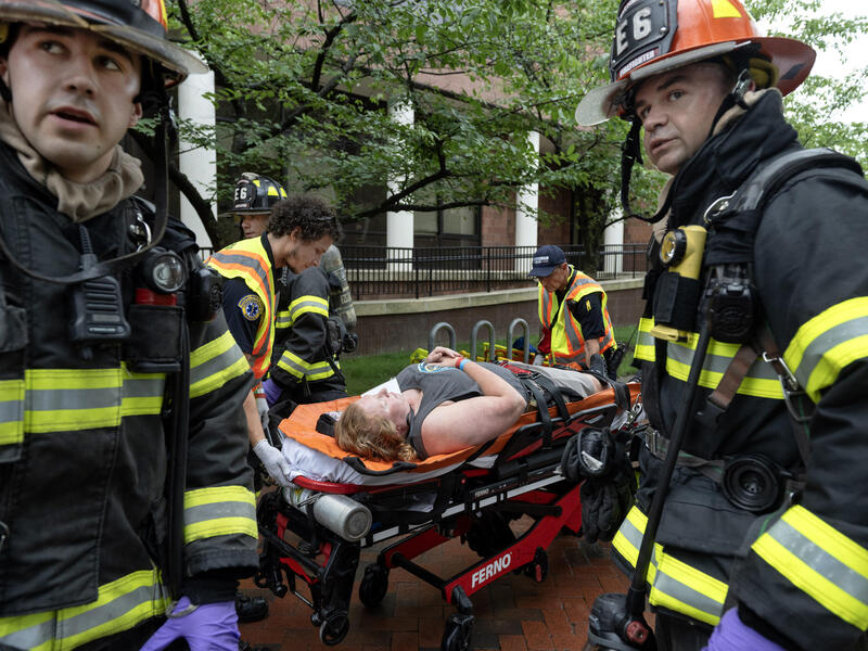 A photo of two firefighters standing in front of a stretcher with a woman laying on it. Two EMS people are rolling the stretcher, while a third firefighter walks past the stretcher. 