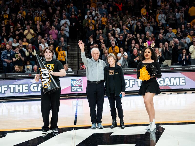 A photo of a man standing to the left of a woman with the arm closest to her behind her back and the other stretched up in the air waving. To the man's left is a another man holding a triangle flag that says \"VCU\" and to the woman's right is a another woman holding pompoms. Behind them, the stadium's seats are filled with people watching. 