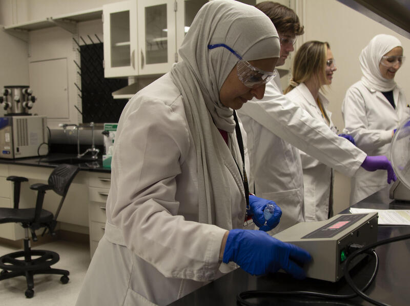 A photo of four people standing in a lab at a counter. They are all wearing white lab coats, safety glasses, and latex gloves. 