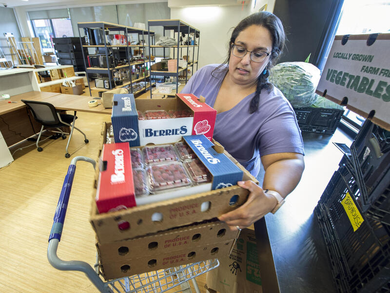 A photo of a woman lifting a box full of raspberries out of a cart. Behind her are shelves with food, and next to her is a metal table with black plastic crates and a white box that says \"VEGTABLES\" in green letters. 