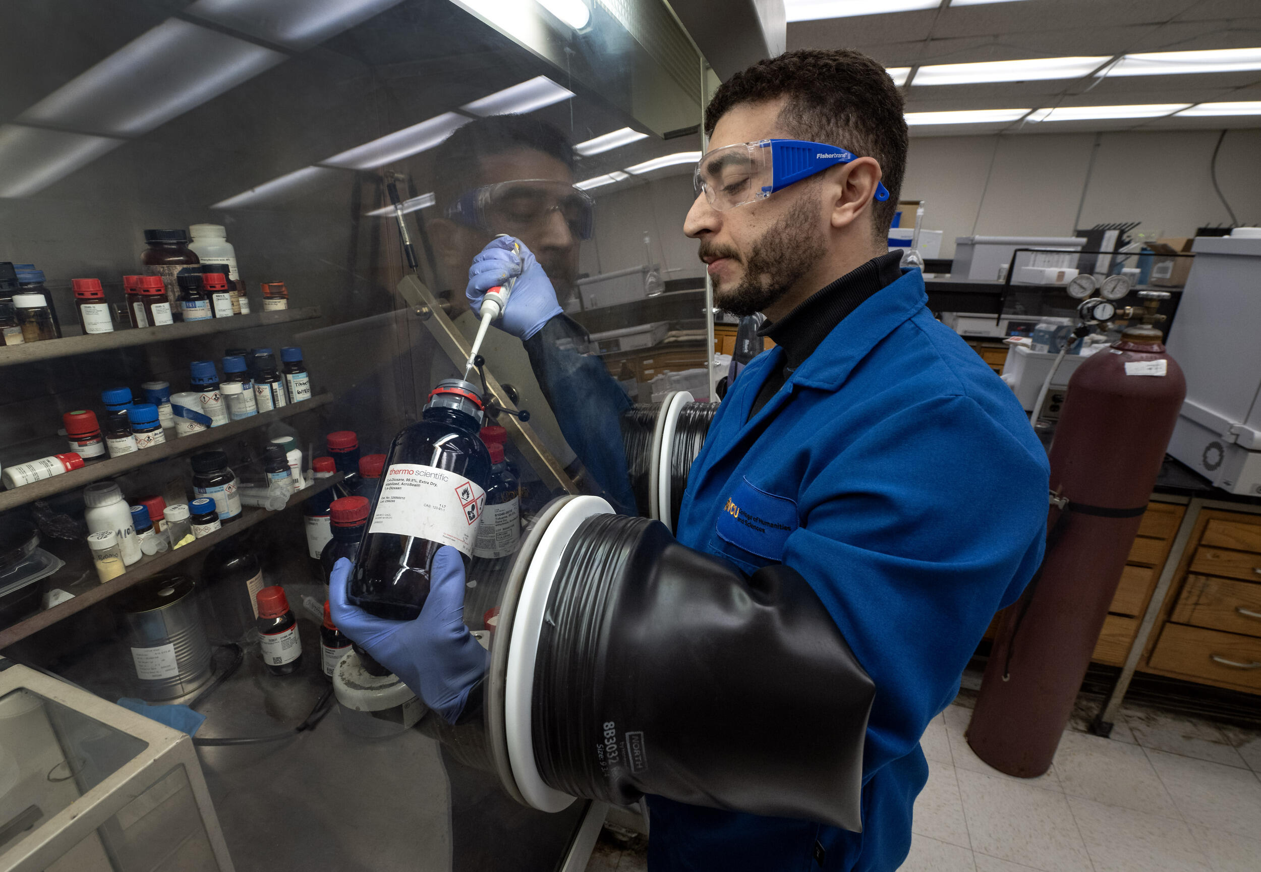 A photo of a man in a science lab using gloves that are attached to a clear wall. On the other side of the wall, his hands are holding a glass jar and a dropper which is extracting liquid from the jar. 