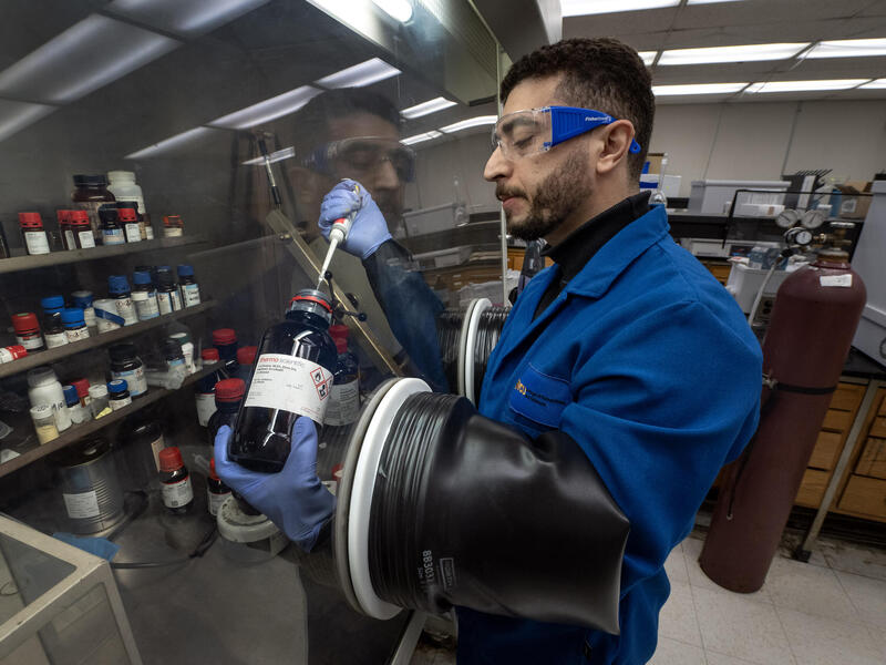 A photo of a man in a science lab using gloves that are attached to a clear wall. On the other side of the wall, his hands are holding a glass jar and a dropper which is extracting liquid from the jar. 