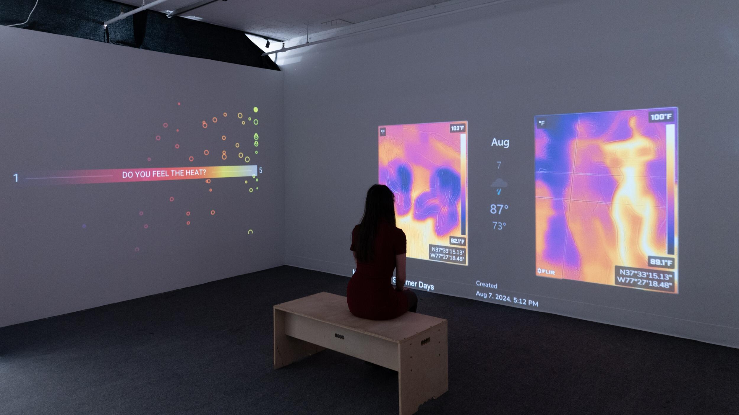 A photo of a woman sitting on a bench in the middle of a dark room. Projections of heat maps are shown on the walls of the room. 