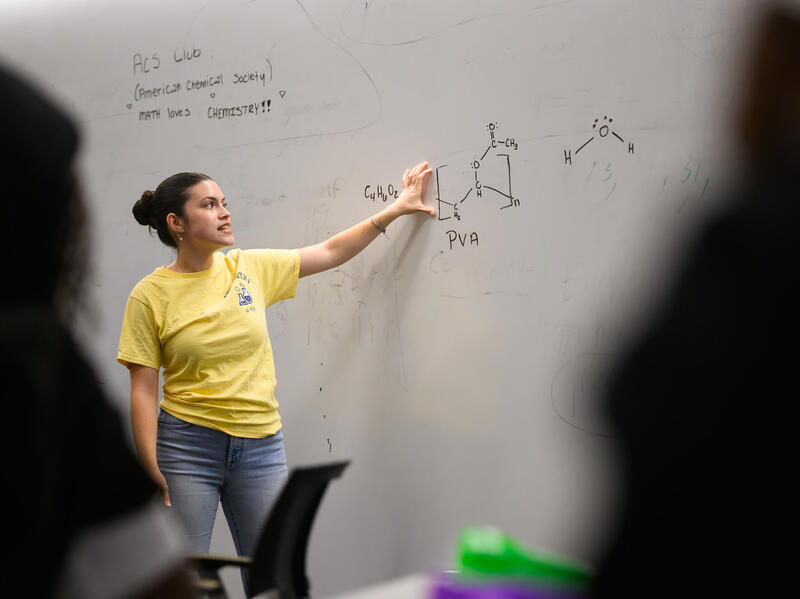 A photo of a woman pointing to a white board. 