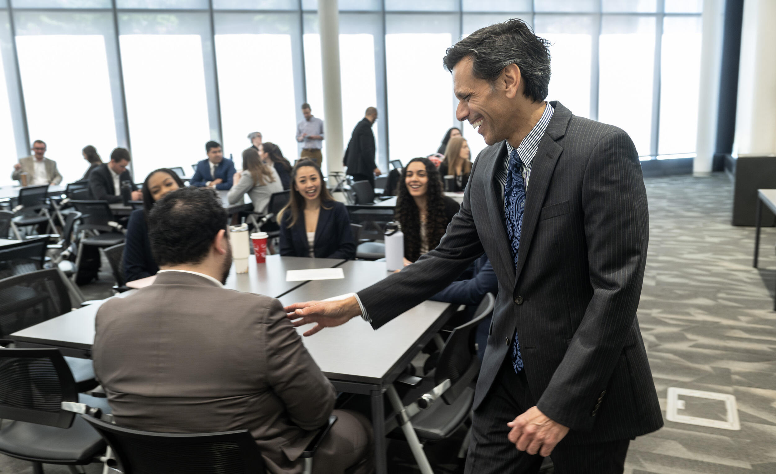 A photo of a man reaching out to another man who is sitting down. There are five people sitting at a table and they are all smaling up at the man who is standing. 
