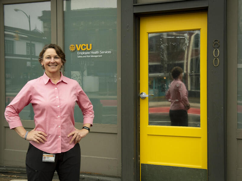 A photo of a woman standing next to the entrance of a building. She has her hands on her hips and is smiling. 