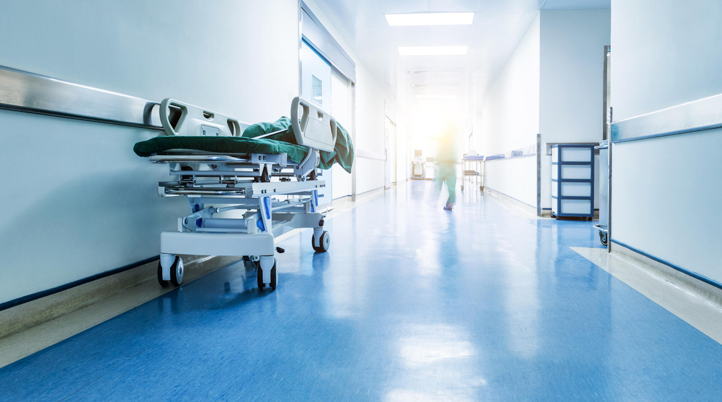 A photo of the hallway of a hospital with a stretcher sitting against the left side wall. Down the hallways is a figure walking who is distorted by a beam of light at the end of the hall. 