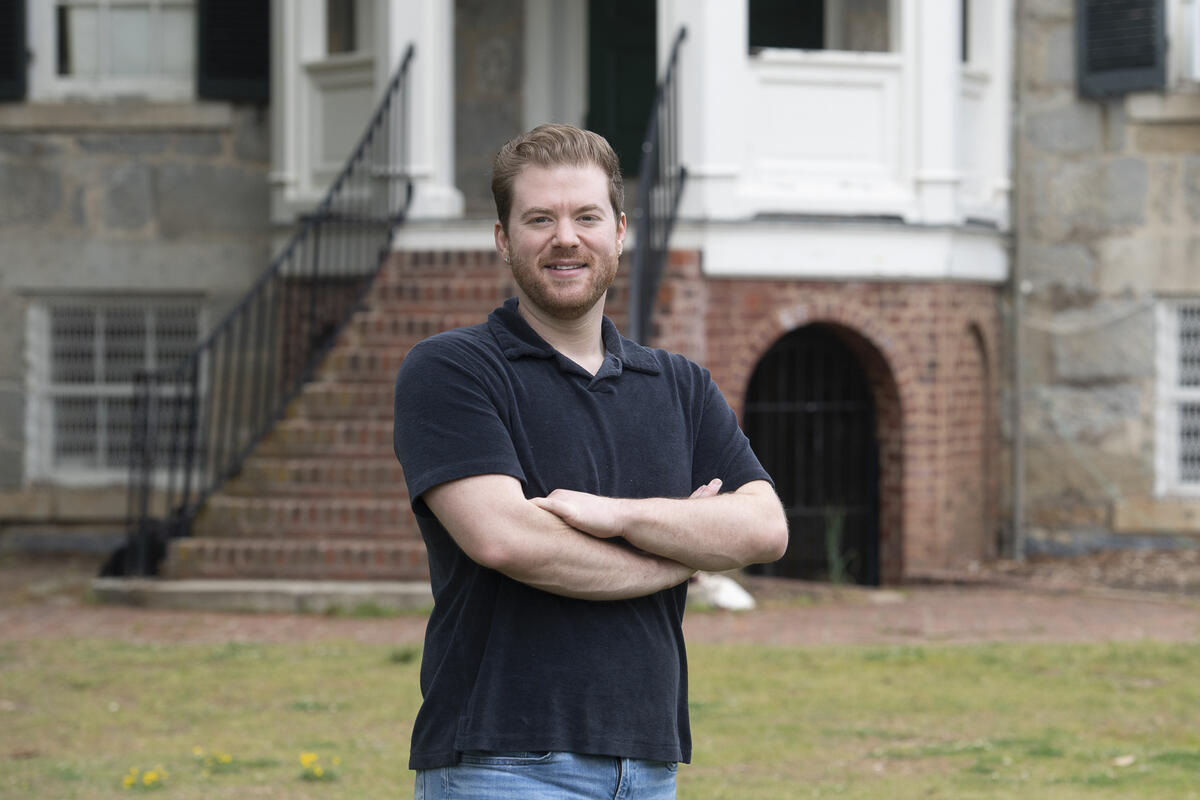A photo of a man from the waist up standing in front of a building and staircase. His arms are crossed against his chest.