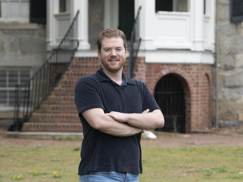 A photo of a man from the waist up standing in front of a building and staircase. His arms are crossed against his chest.