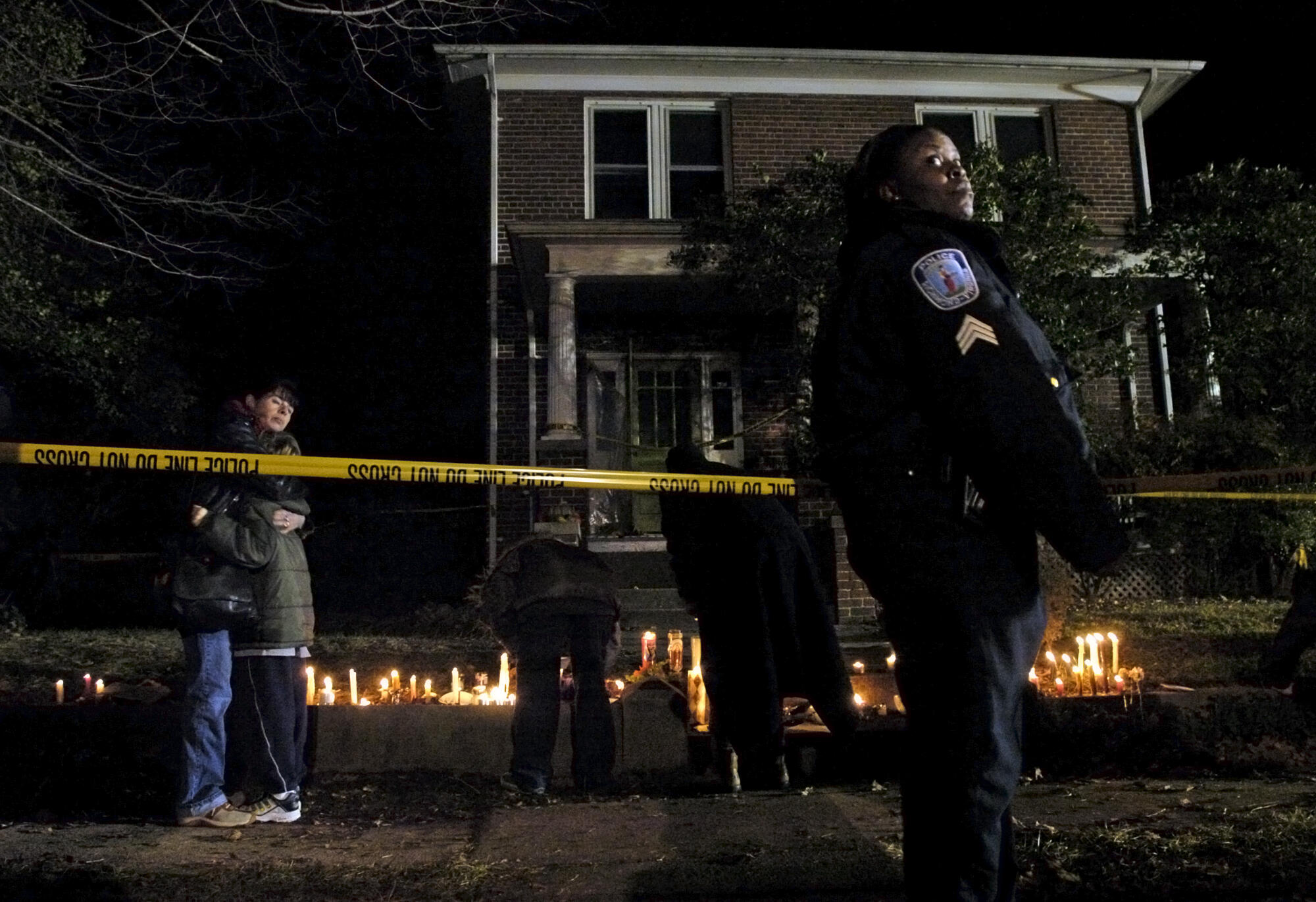 A police officer standing in front of a yellow cation tape in front of a home. Behidn the tape people standing in front of candles. 