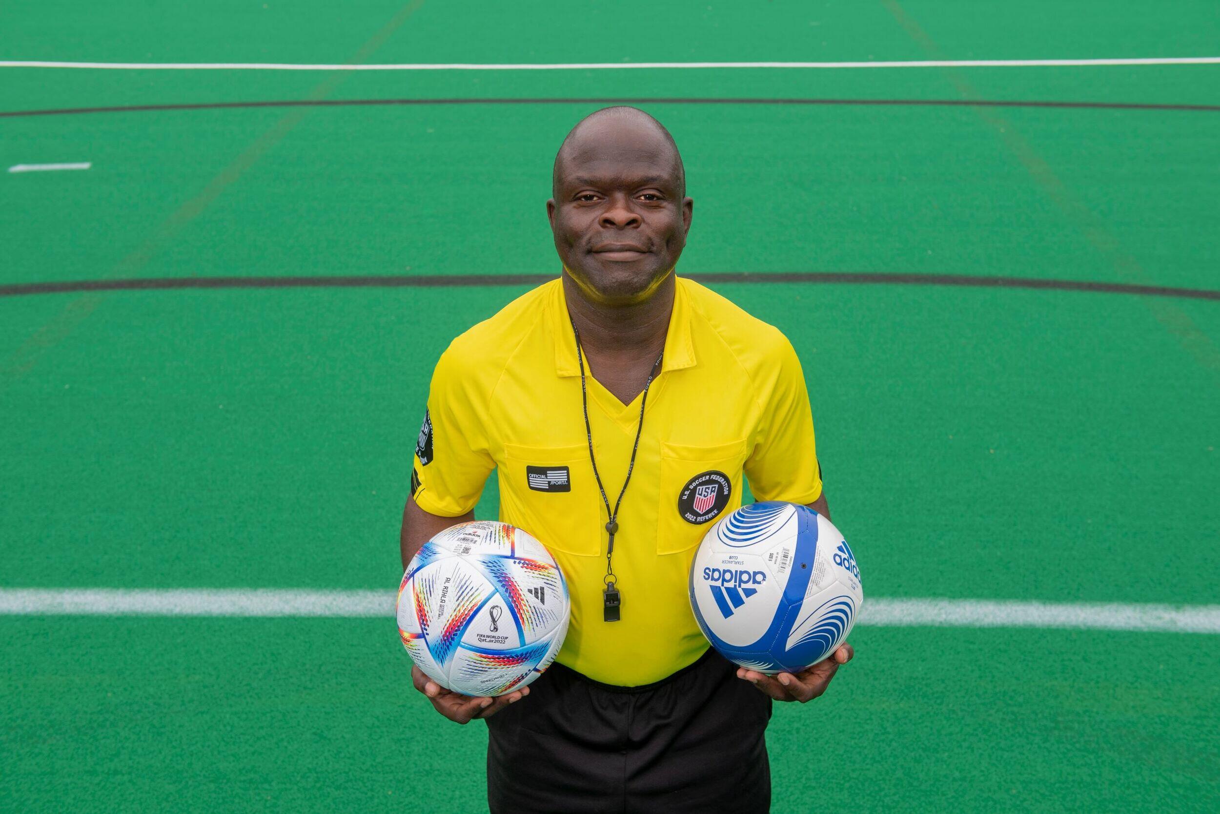 Kwasi Deitutu, wearing a soccer ref's uniform, holds two soccer balls.