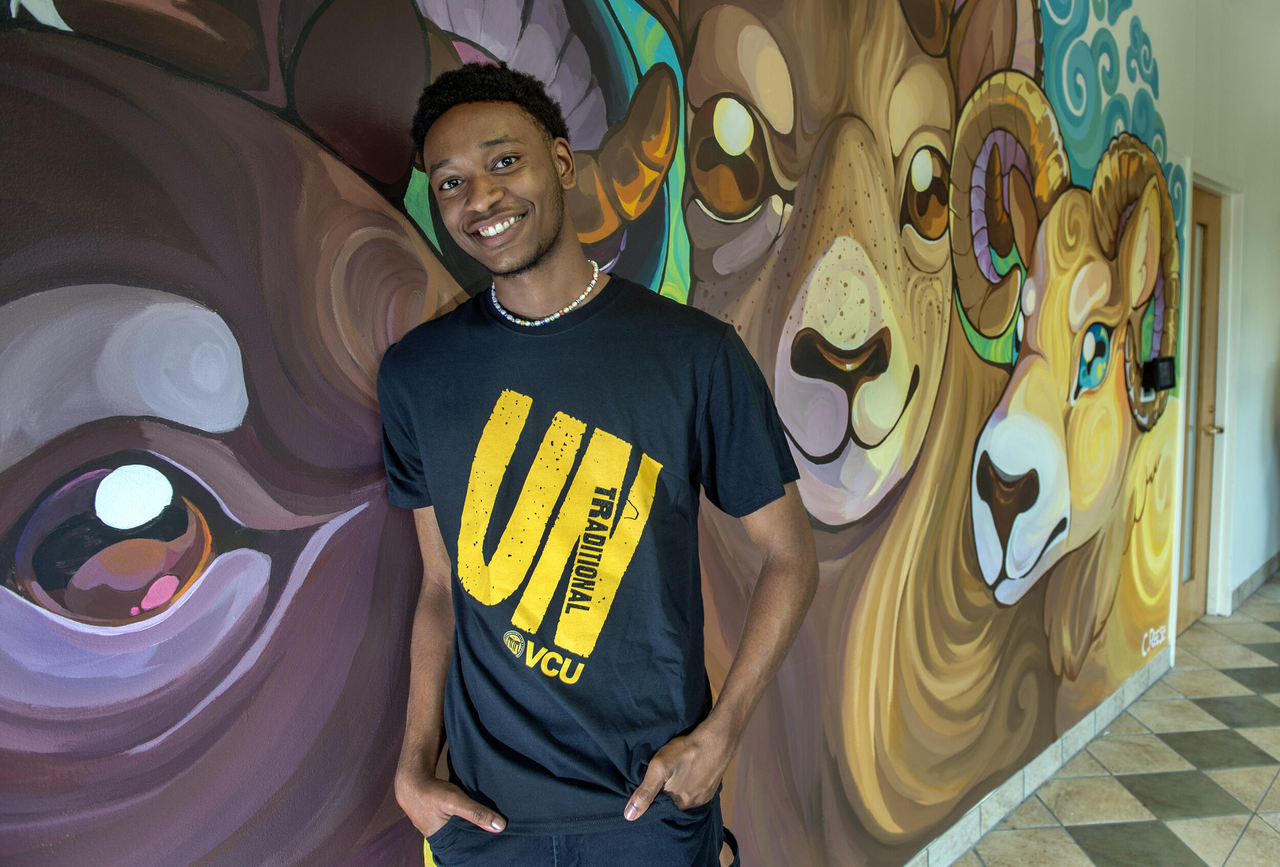 A photo of a man from the waist up leaning against a wall with a mural of three rams on it. The man is wearing a black shirt with yellow letters on it. The text on his shirt says \"UN TRADITIONAL\" and \"VCU.\"