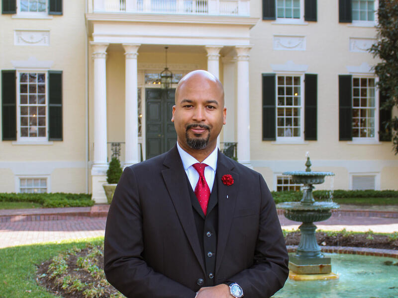 A photo of a man in a suit and tie standing in front of Virginia’s Executive Mansion.