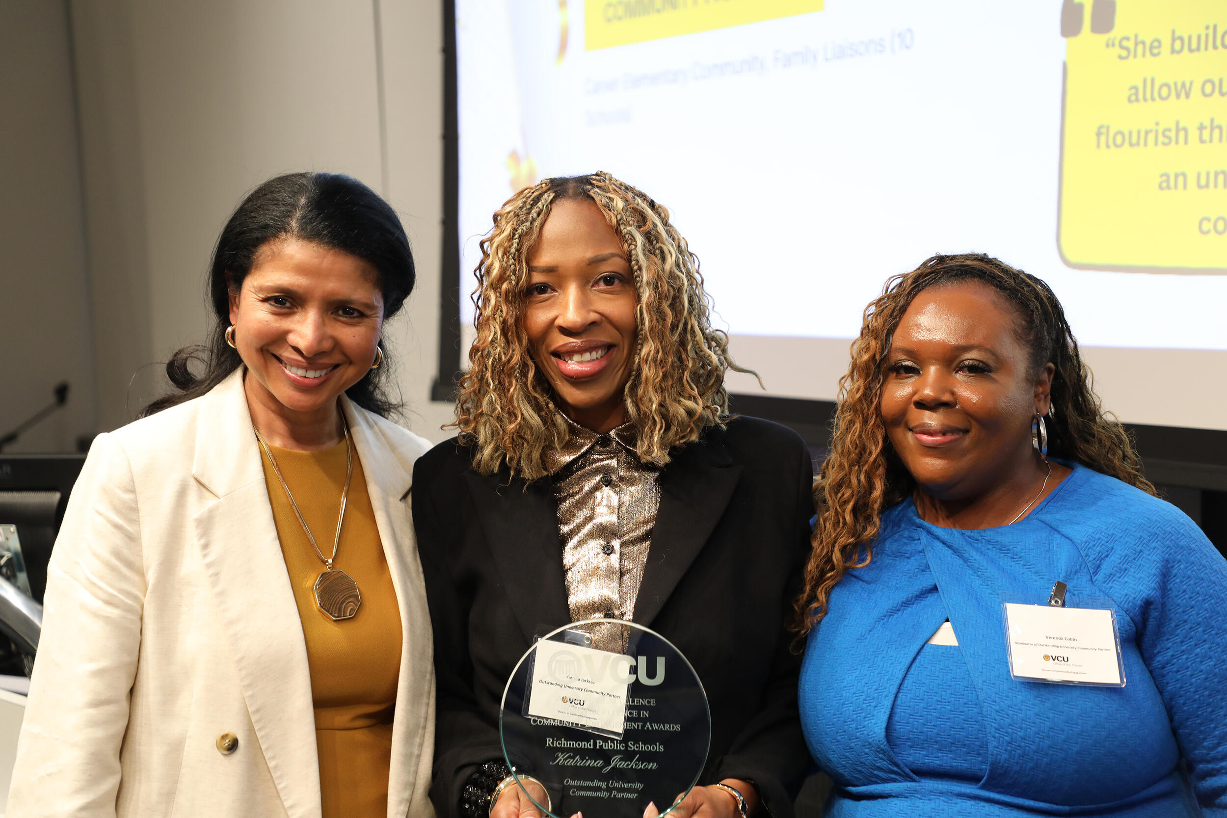 A photo of three woman from the chest up. The woman in the center is holding a glass award trophy. 