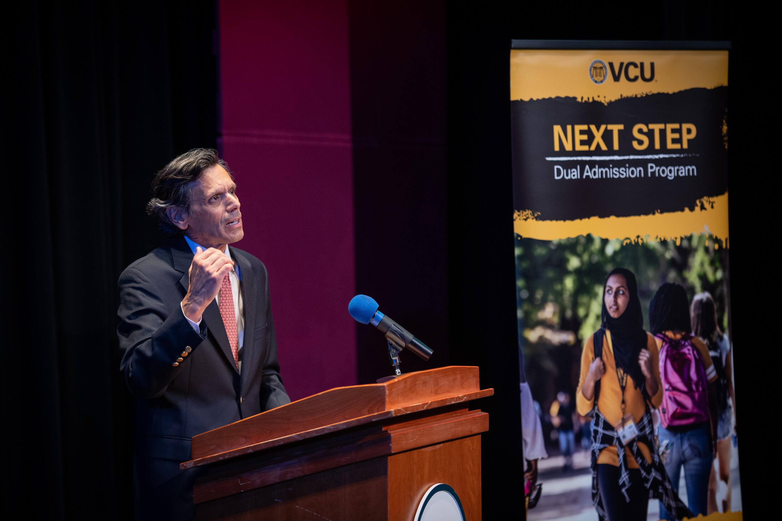 A man in a coat and tie gestures while speaking at a lectern into a microphone.